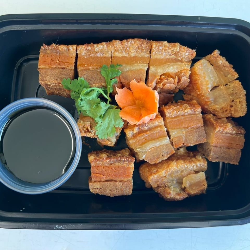 Crispy pork belly pieces with cilantro, a carrot flower, and a side of dark sauce in a black container.