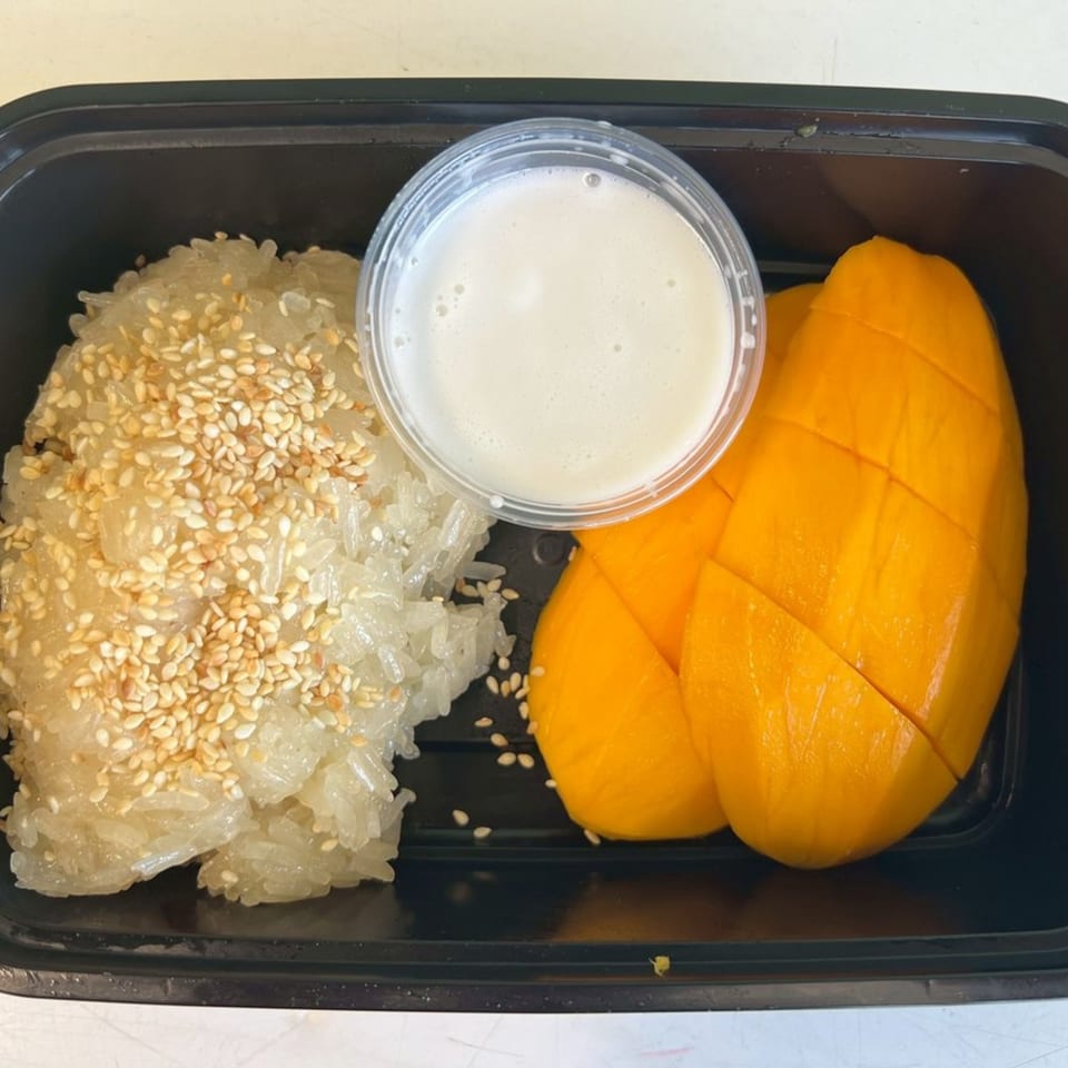 Thai mango sticky rice with sesame seeds and a side of coconut milk in a black container.