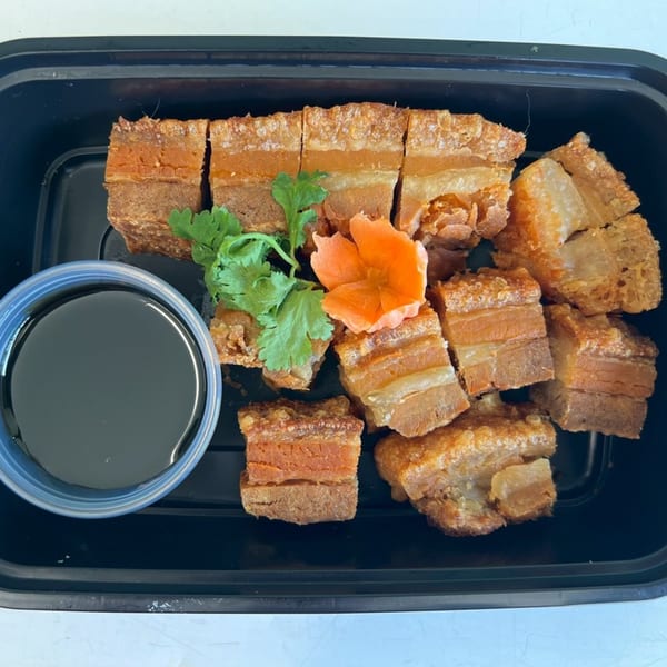 Crispy pork belly pieces with cilantro, a carrot flower, and a side of dark sauce in a black container.