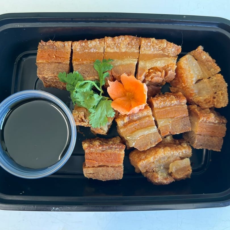 Crispy pork belly pieces with cilantro, a carrot flower, and a side of dark sauce in a black container.