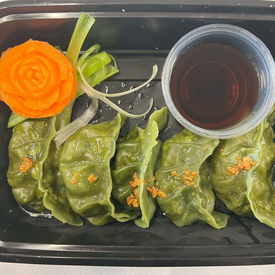 Green dumplings with orange garnish and a side of dipping sauce in a black tray.