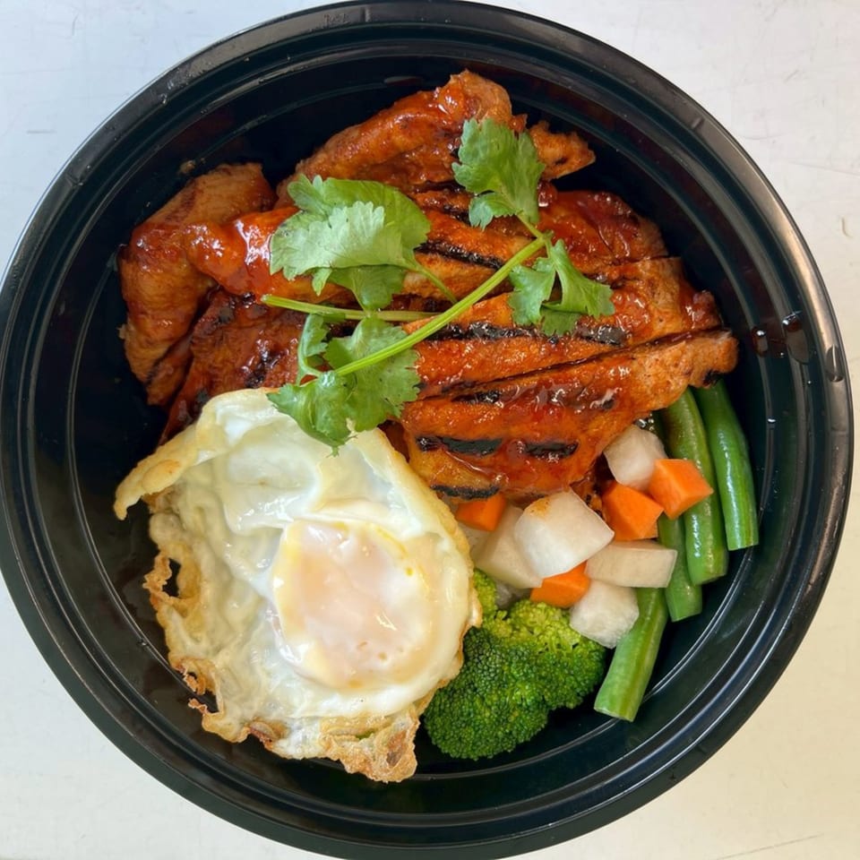 Thai Accent's grilled pork with a fried egg, broccoli, carrots, and green beans in a black bowl.